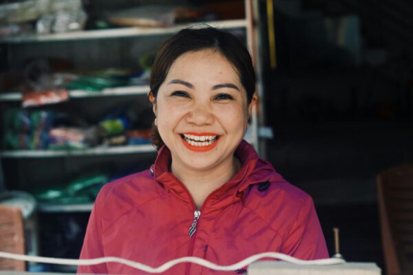 A cheerful woman in a pink jacket smiling broadly, standing in front of a market stall.