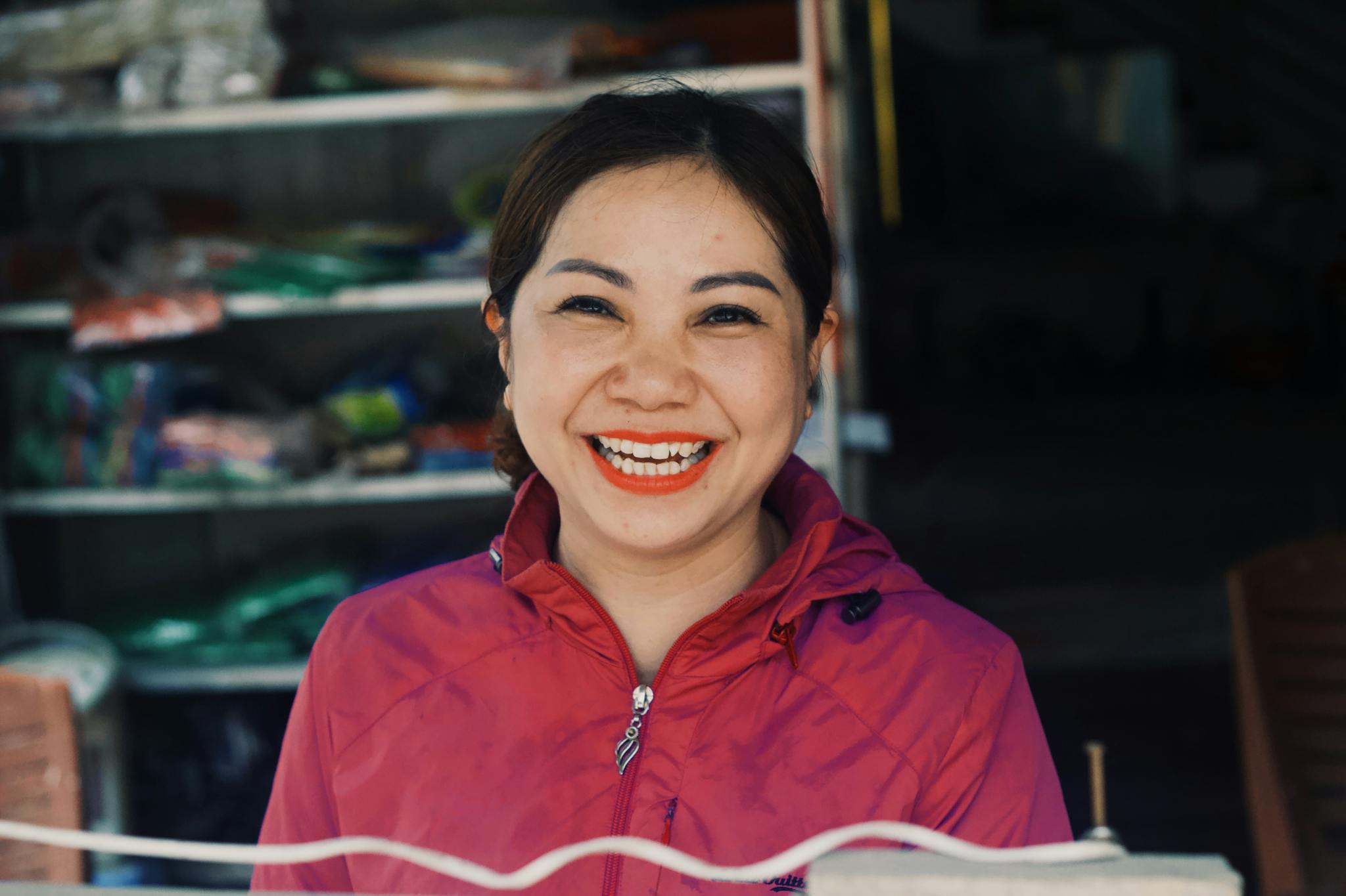 A cheerful woman in a pink jacket smiling broadly, standing in front of a market stall.